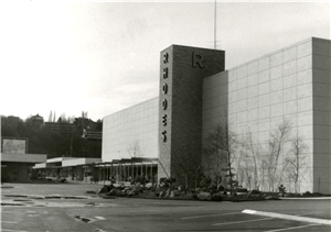 A tall mid century shopping center with an empty parking lot. On the front of the building is a large vertical sign that says Rhodes. 